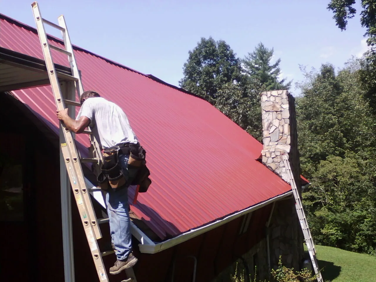 Roofer on a ladder beside a red metal roof and stone chimney — Soffit & Fascia Repair by Roofing Brothers in Longport